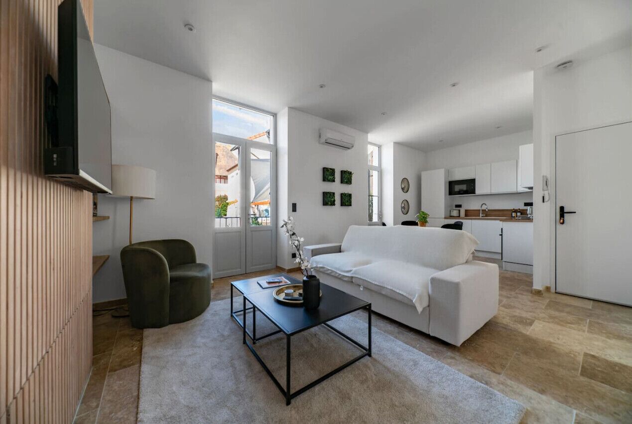 Ein modernes Wohnzimmer mit einem weißen Sofa, einem schwarzen Couchtisch und einem grünen Sessel. Im Hintergrund ist eine Küchenzeile erkennbar.