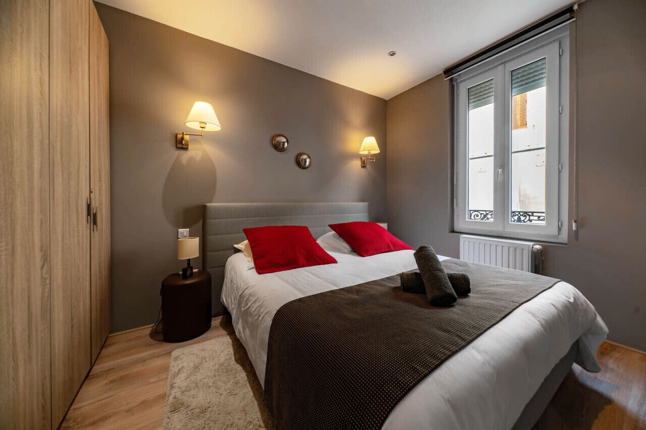 A bedroom with a bed, two bedside lamps, a window with blinds, and a radiator.