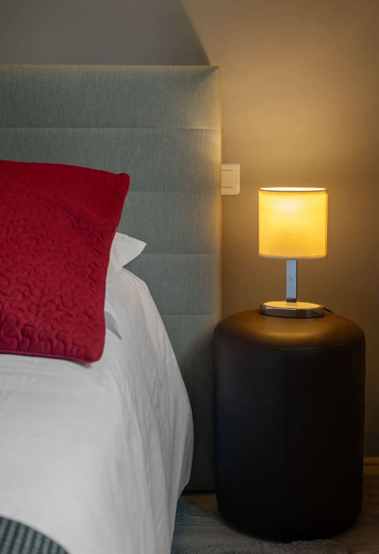 A neatly made bed with a red pillow, a bedside lamp, and a grey headboard.