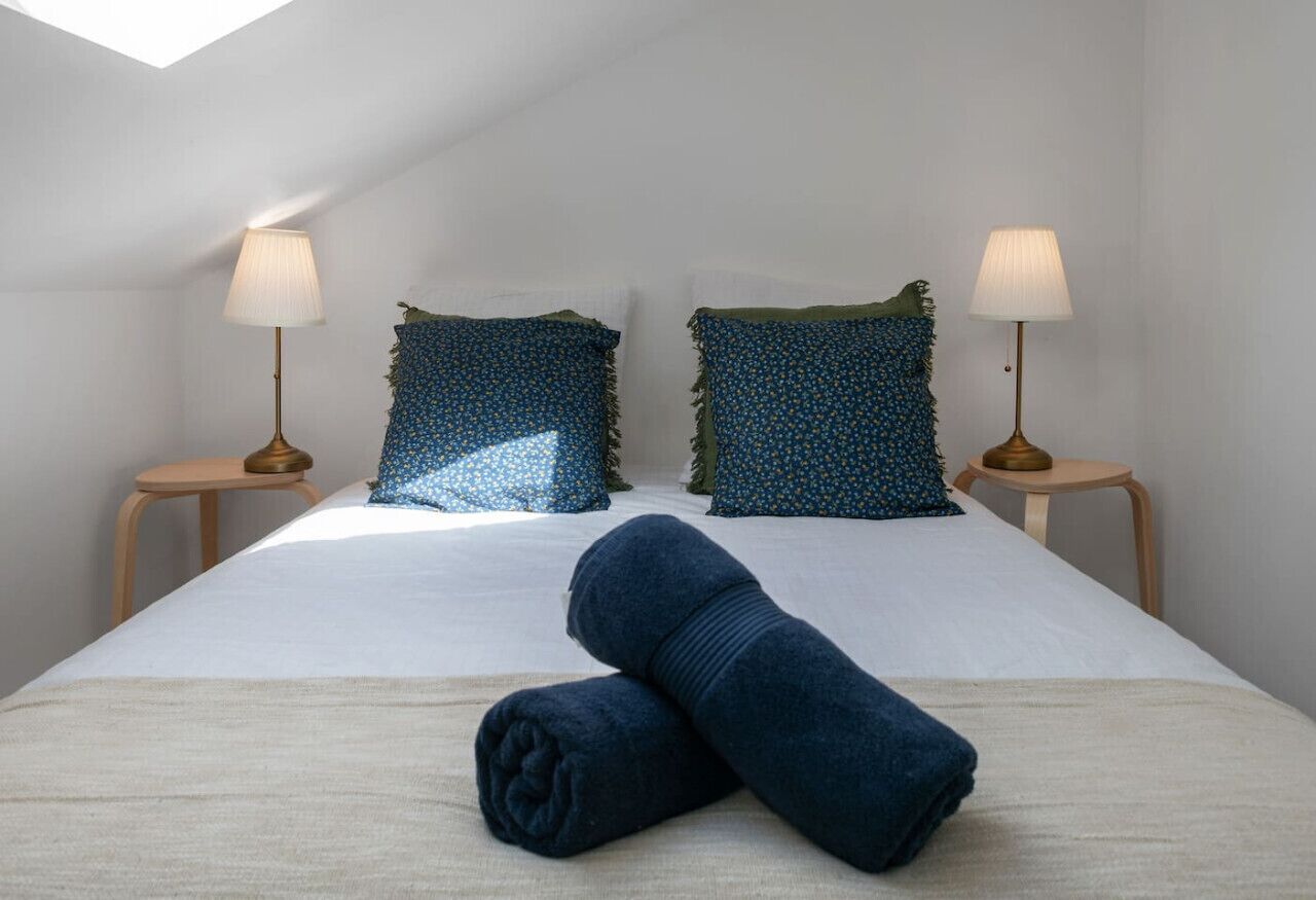 Una cama bien tendida con dos almohadas, dos lámparas de noche y una toalla enrollada sobre la cama.