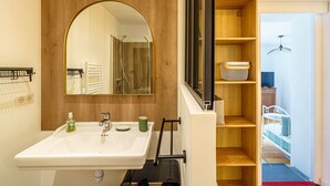 Une salle de bain avec un grand miroir, un lavabo et une douche. On y trouve une étagère en bois avec un panier et un luminaire suspendu.