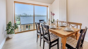 A dining area with a large table set for four, surrounded by chairs with cushioned seats. There is a potted plant on the left and a vase with flowers on the table. The room has large windows offering a view of a body of water and trees. - Hollyhocks - UK47975 (Woodford)