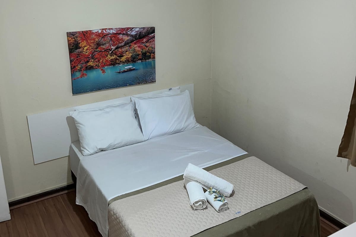 A neatly made bed with a white headboard, a ceiling fan, and a painting of a scenic landscape on the wall.