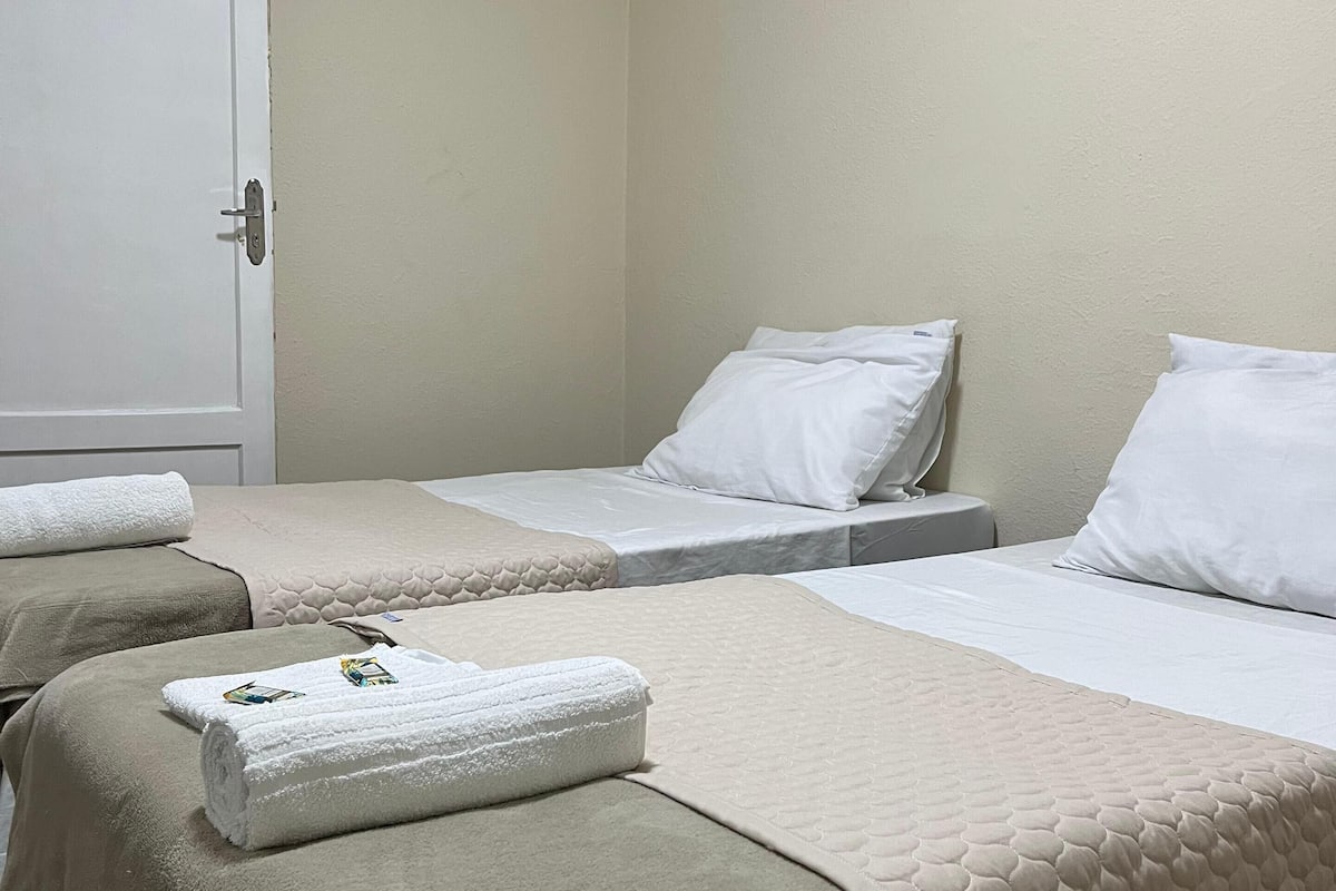 A hotel room with two single beds, each with a white pillow and a beige quilted blanket. There is a white door with a handle, and a white towel is placed on the floor beside one of the beds.