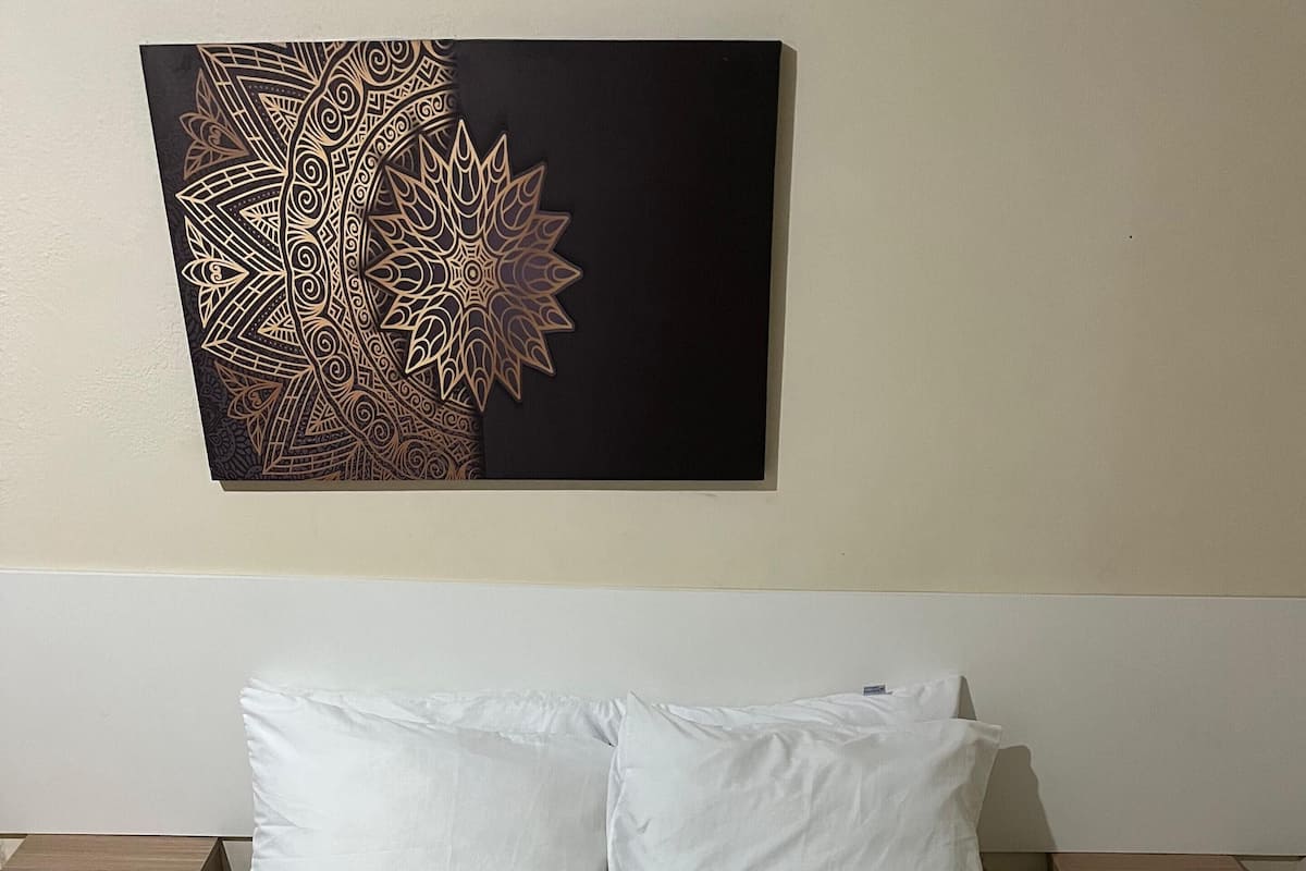 A neatly made bed with white linens, a beige quilted mattress cover, and two white pillows. There are two wooden nightstands, one on each side of the bed, each with a drawer. A decorative wall piece is hung above the bed.