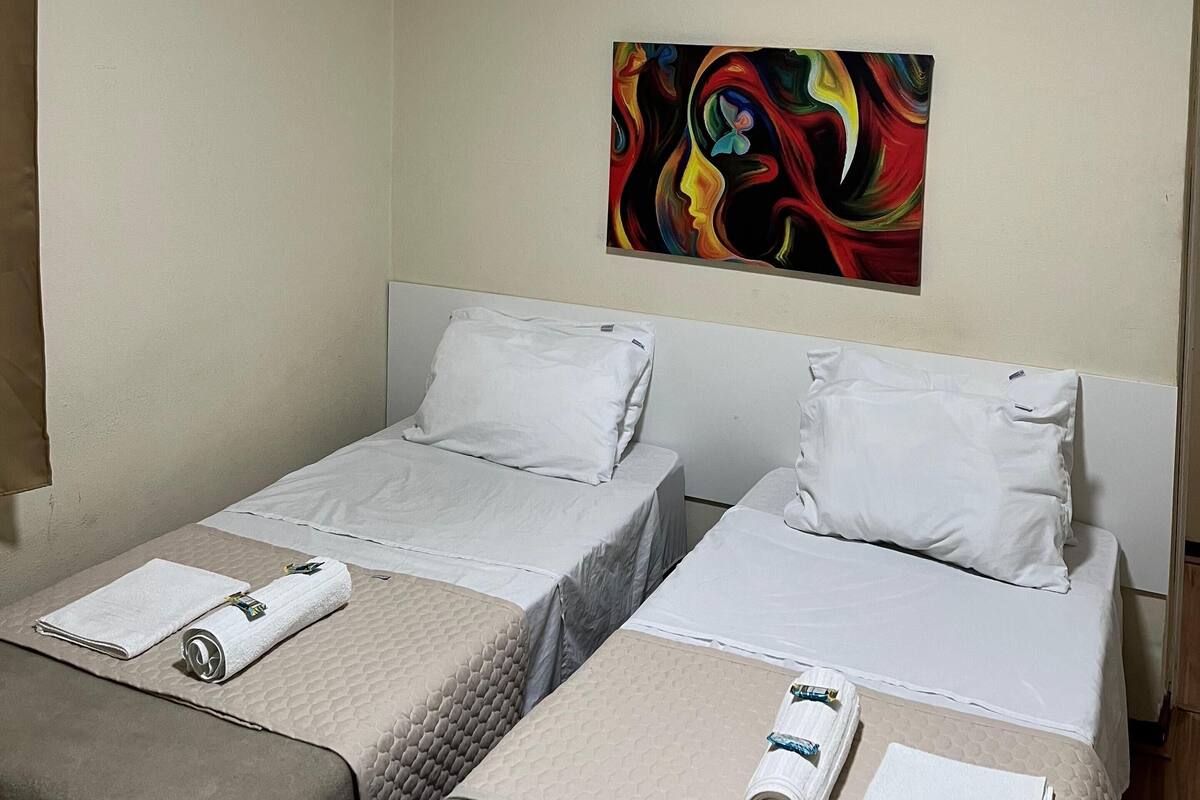 A hotel room with two beds, each with a white pillow and a rolled towel. A colorful abstract painting is on the wall above the beds.