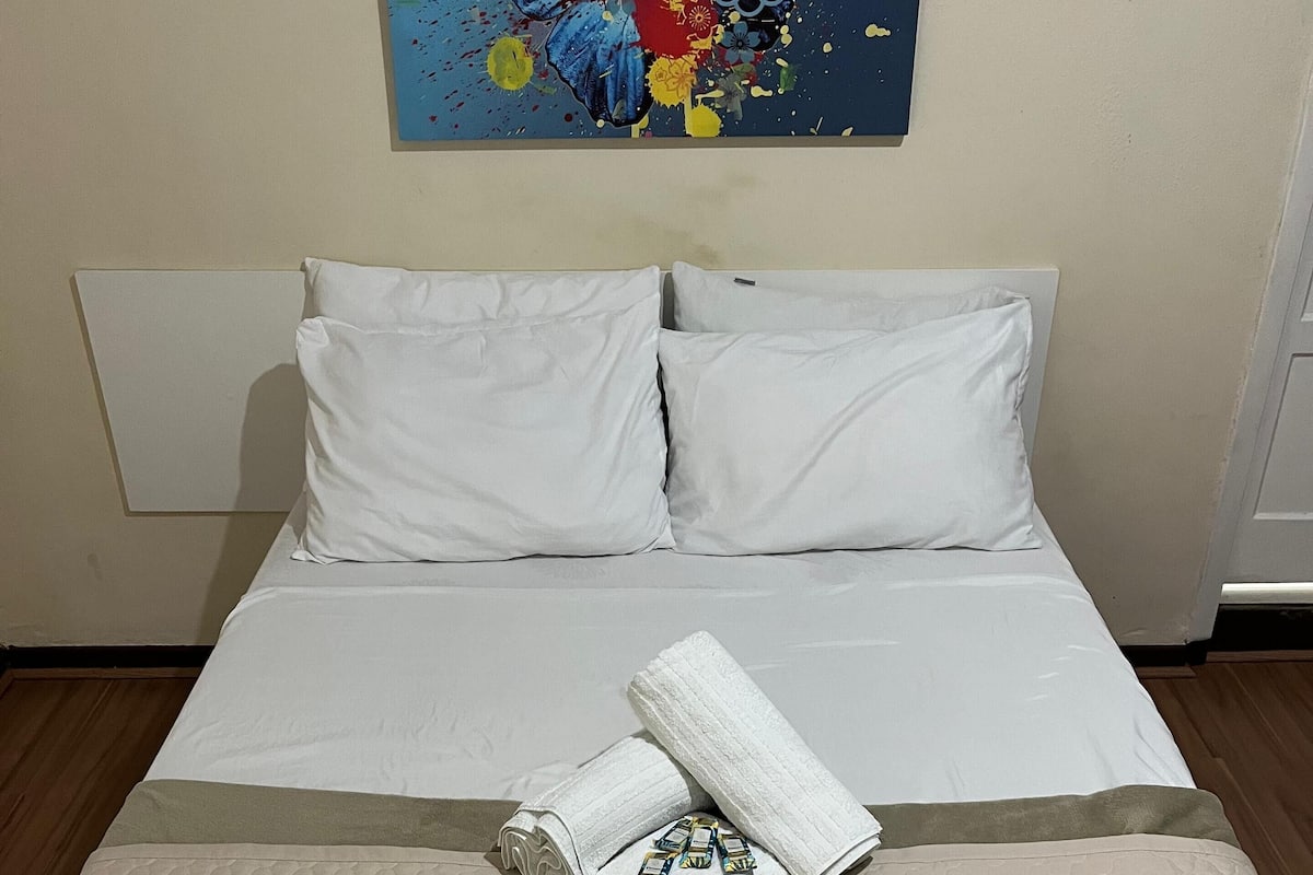 A neatly made bed with white linens and a decorative pillow, a rolled towel, and a pillowcase with a floral pattern on a textured bedspread.