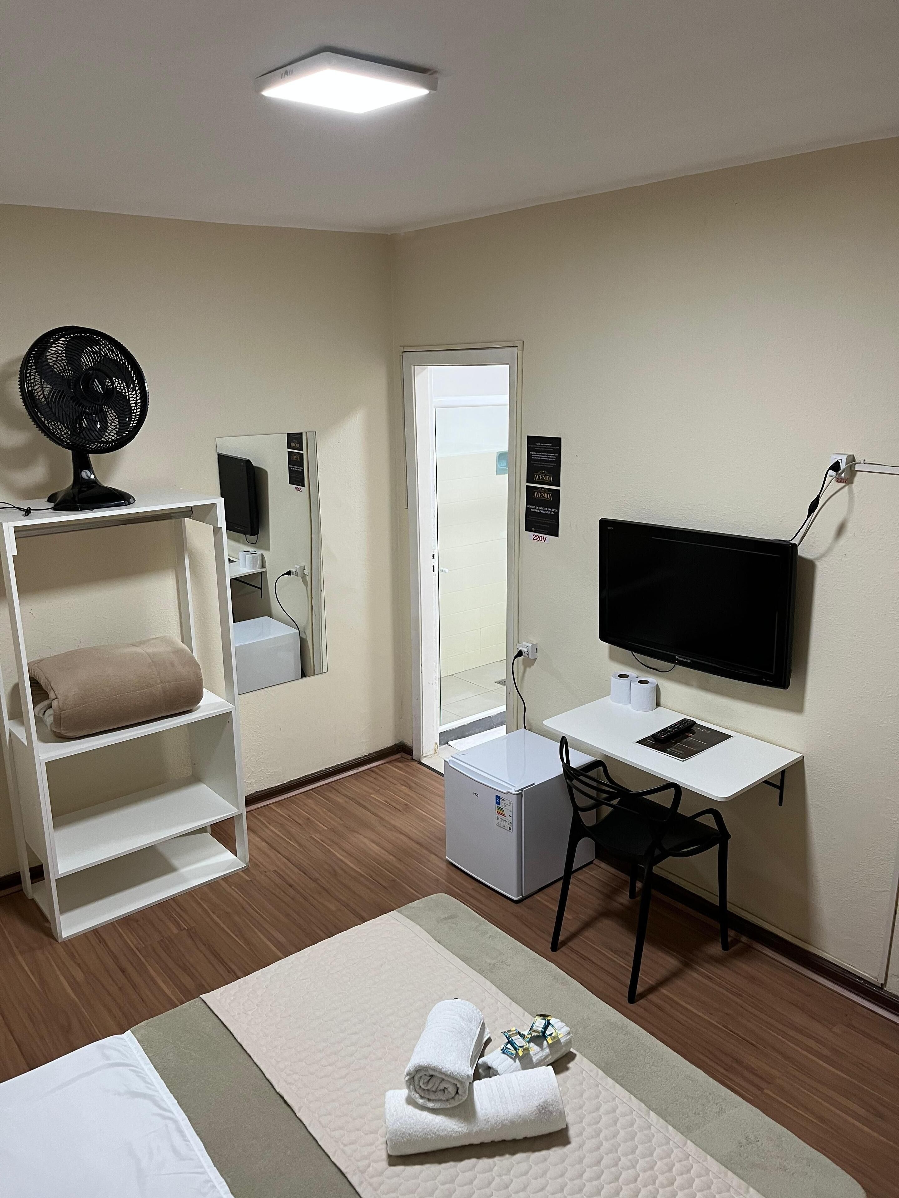 A hotel room with a bed, a desk with a computer, a TV, a fan, and a small refrigerator.