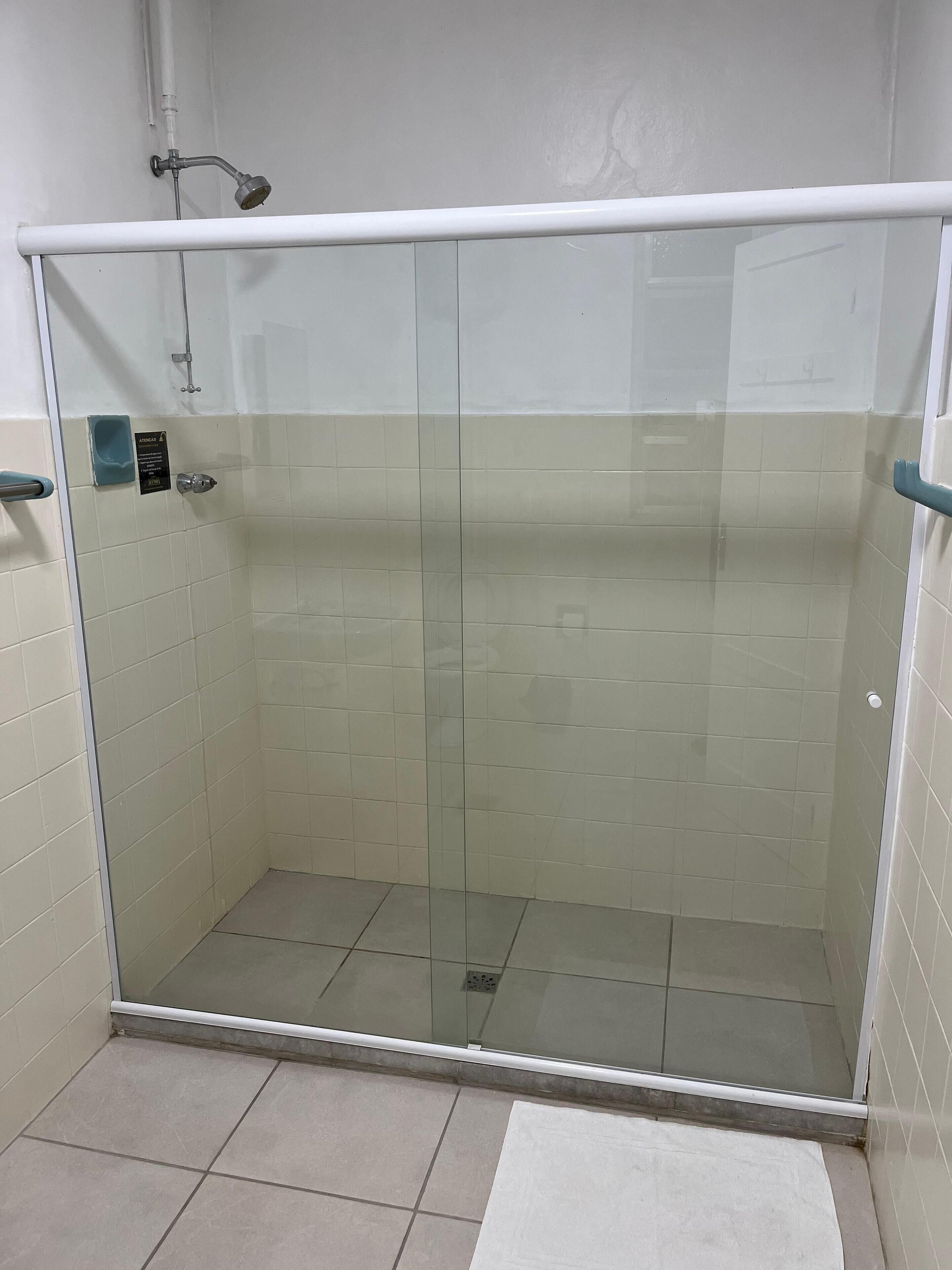 Cabina de ducha con paredes y suelo de baldosas blancas, desagüe visible y pasamanos en el lado derecho.