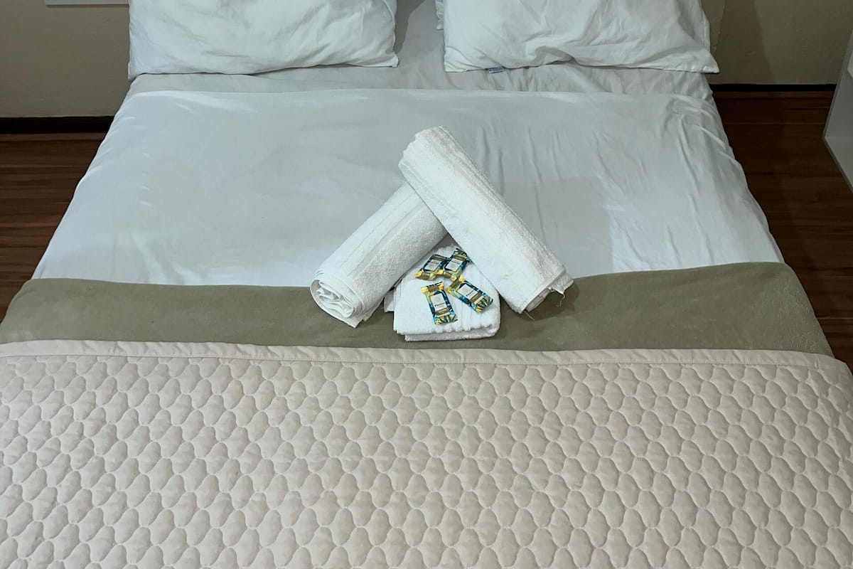 A neatly made bed with white linens, two pillows, and rolled towels on a textured mattress.
