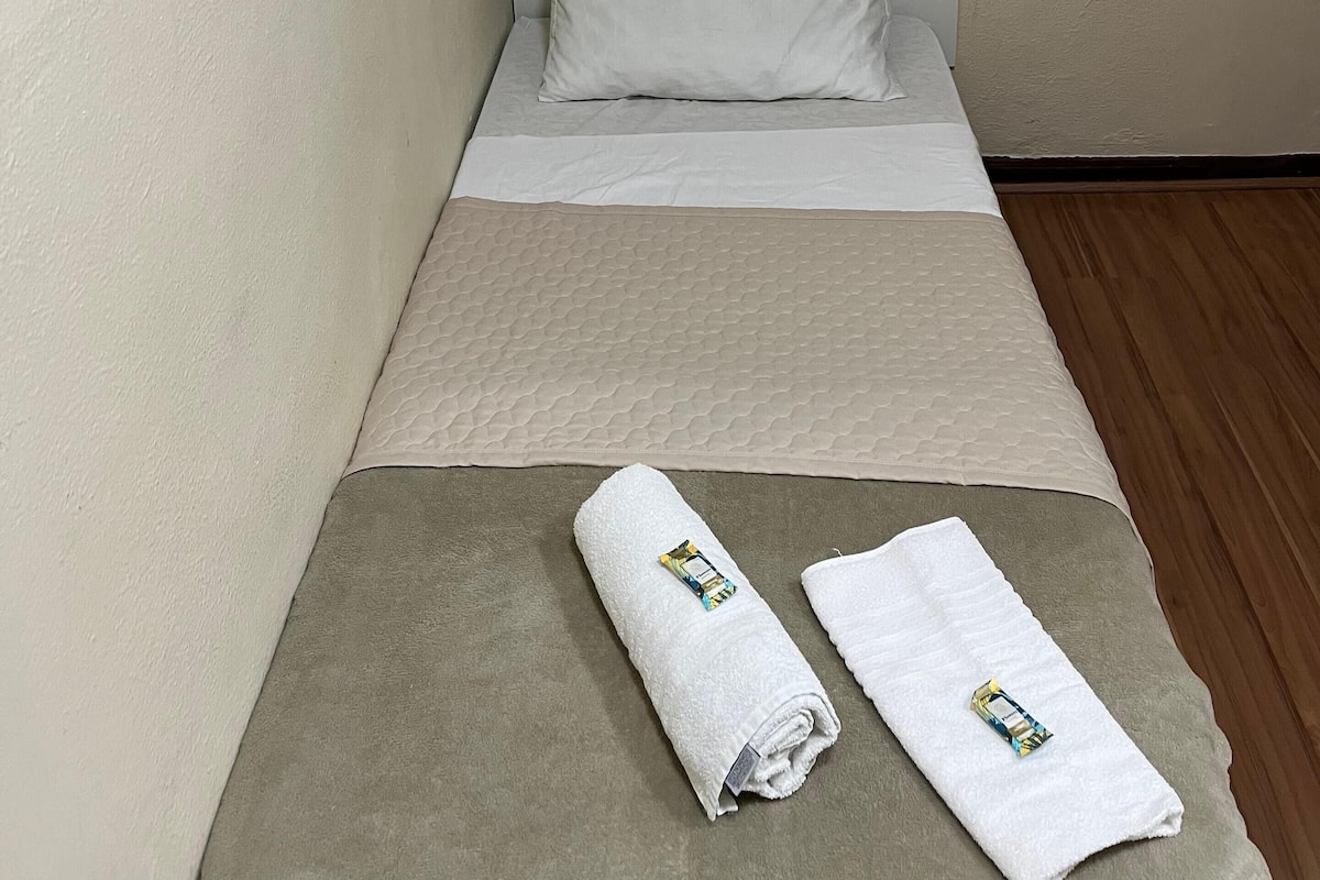 A small, neatly made bed with white linens, a beige blanket, and a towel with a colorful tag. A painting with blue and red tones is hung on the wall above the bed.