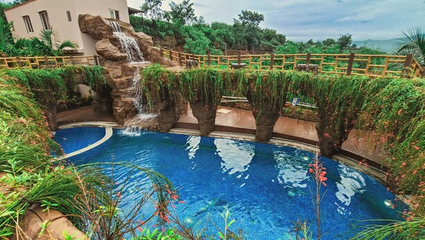 A swimming pool with a waterfall feature, surrounded by lush greenery and a wooden fence. - Regenta Bharti Amusement & Adventure Resort Pune, Urulikanchan (Daund)
