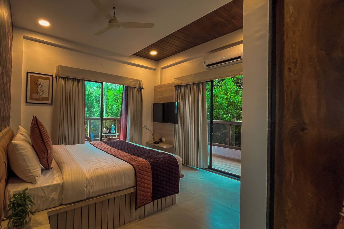 A bedroom with a bed, curtains, a TV, and a view of greenery outside.