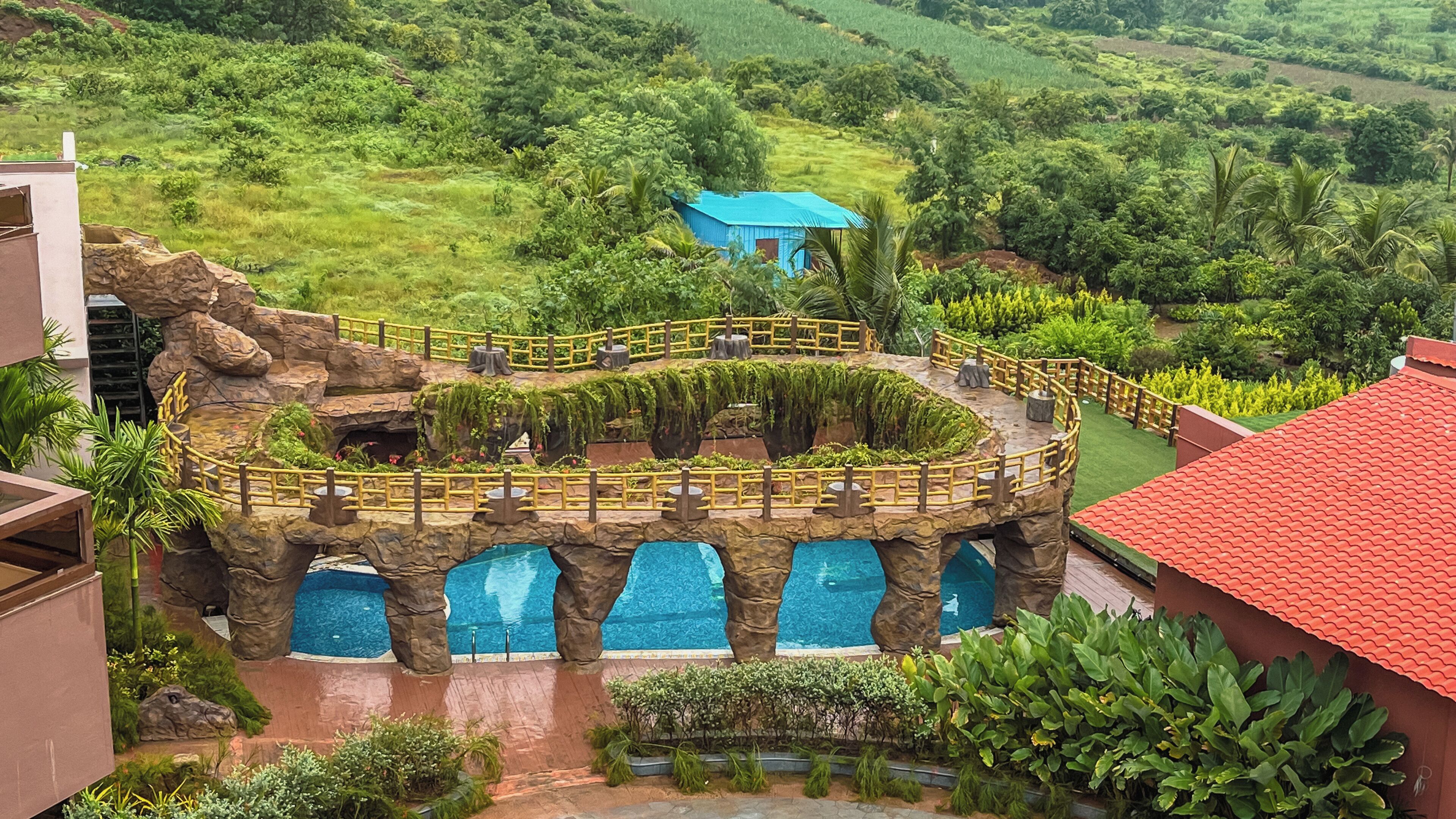 Un resort con una piscina circular rodeada por un puente rocoso, abundante vegetación y un edificio azul a lo lejos.