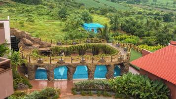 Un resort con una piscina circular rodeada por un puente rocoso, abundante vegetación y un edificio azul a lo lejos.