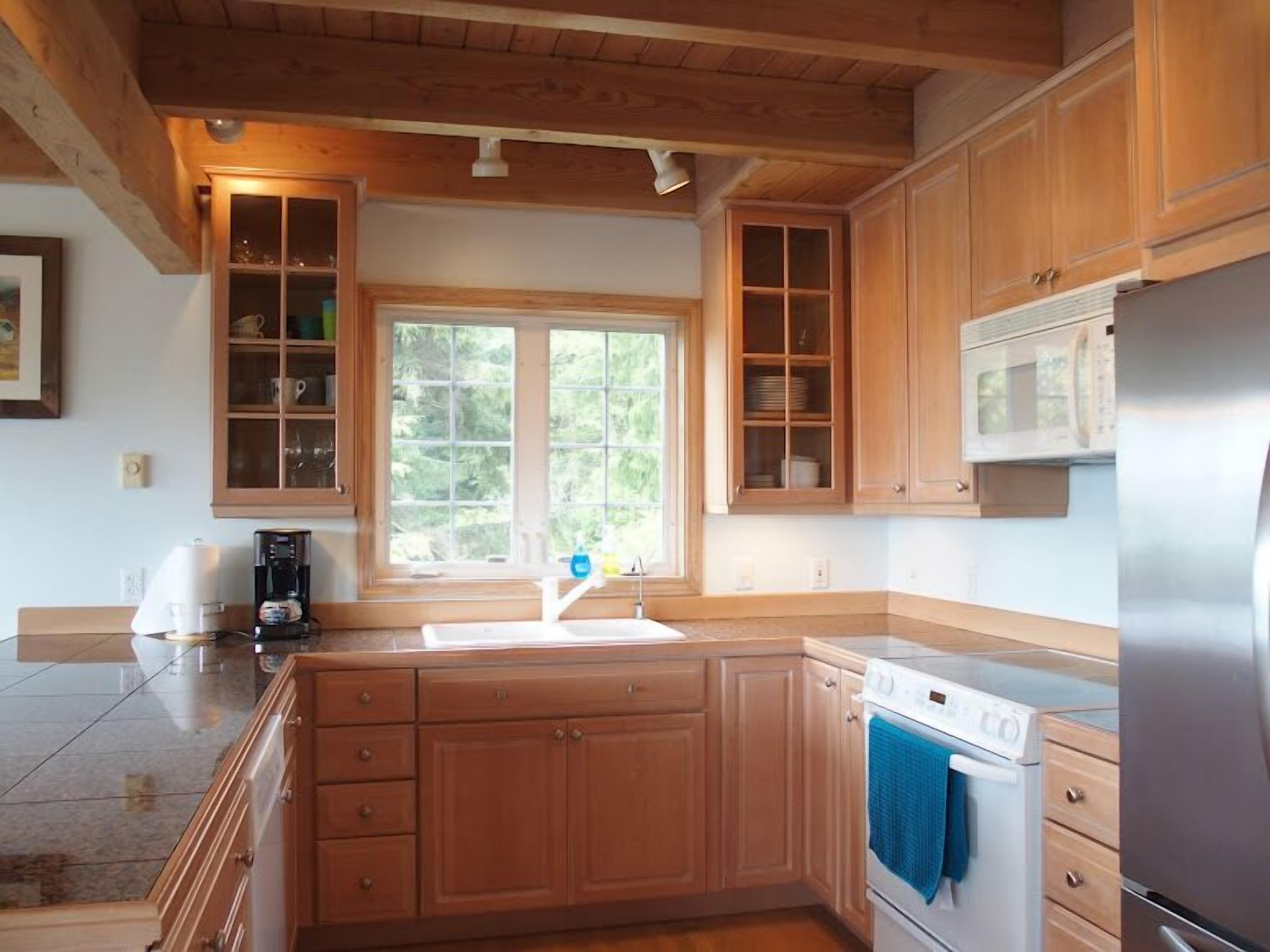 A kitchen with wooden cabinets, a stainless steel refrigerator, a microwave, and a window with a view of greenery.