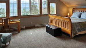 Une chambre avec un plafond en pente, un ventilateur de plafond, un lit recouvert d’une couvre-lit à motifs, un bureau avec une chaise et de grandes fenêtres donnant sur les arbres et le ciel.