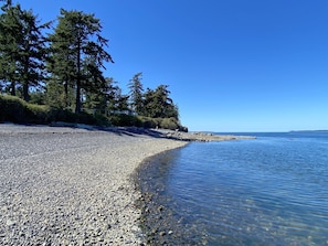 Una playa de guijarros con agua transparente, árboles y una costa lejana.