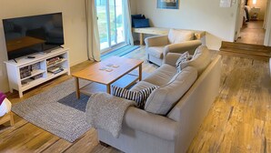 A living room with a TV, sofa, coffee table, and a view of the outdoors through a large window. - North Shore Retreat: Private Beach Access, Tennis/Pickleball Court, Pets Welcome (Eastsound)