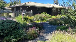 A house with a dark exterior, a metal roof, and a gravel driveway leading to a wooden fence. - North Shore Retreat: Private Beach Access, Tennis/Pickleball Court, Pets Welcome (Eastsound)