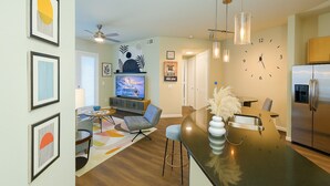 A modern kitchen with a central island, a dining area with a table and chairs, and a living room with a TV and seating. - Sunshine Stay | 2BR Galleria Apartment Nr Mall (Houston)