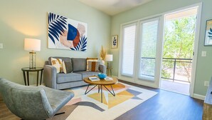 A living room with a gray sofa, a round coffee table, and a gray armchair. There is a large abstract painting on the wall and a window with blinds. - Sunshine Stay | 2BR Galleria Apartment Nr Mall (Houston)