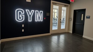 A gym entrance with a neon sign, a door, and a floor with dark tiles. - Sunshine Stay | 2BR Galleria Apartment Nr Mall (Houston)