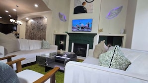 A modern living room with a white sofa, a coffee table, and a fireplace. There is a TV mounted on the wall above the fireplace.
