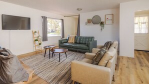 Una sala de estar con un sofá verde, un sillón beige y una mesita redonda. Hay un televisor montado en la pared, una escalera con plantas y una ventana con cortinas.
