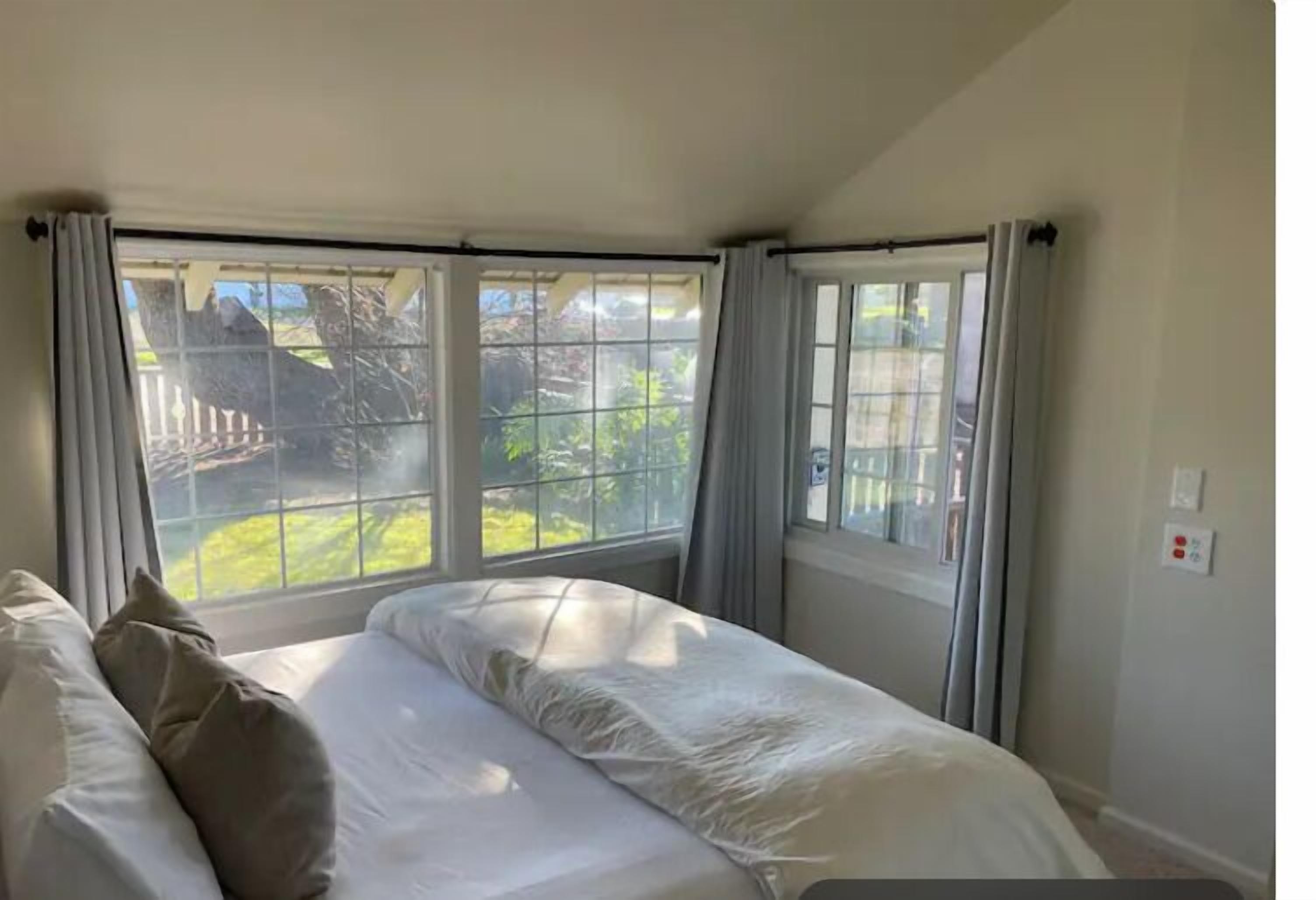 A bedroom with a bed, two windows with curtains, and a view of a garden.