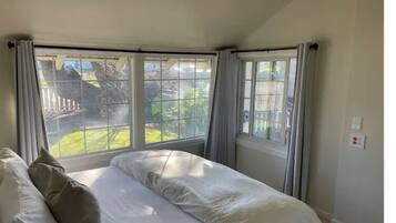 A bedroom with a bed, two windows with curtains, and a view of a garden.