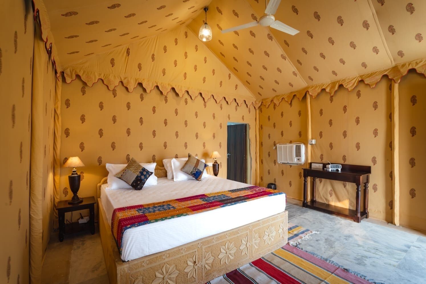 A bedroom with a bed, two bedside lamps, a ceiling fan, an air conditioning unit, and a small table with a radio.