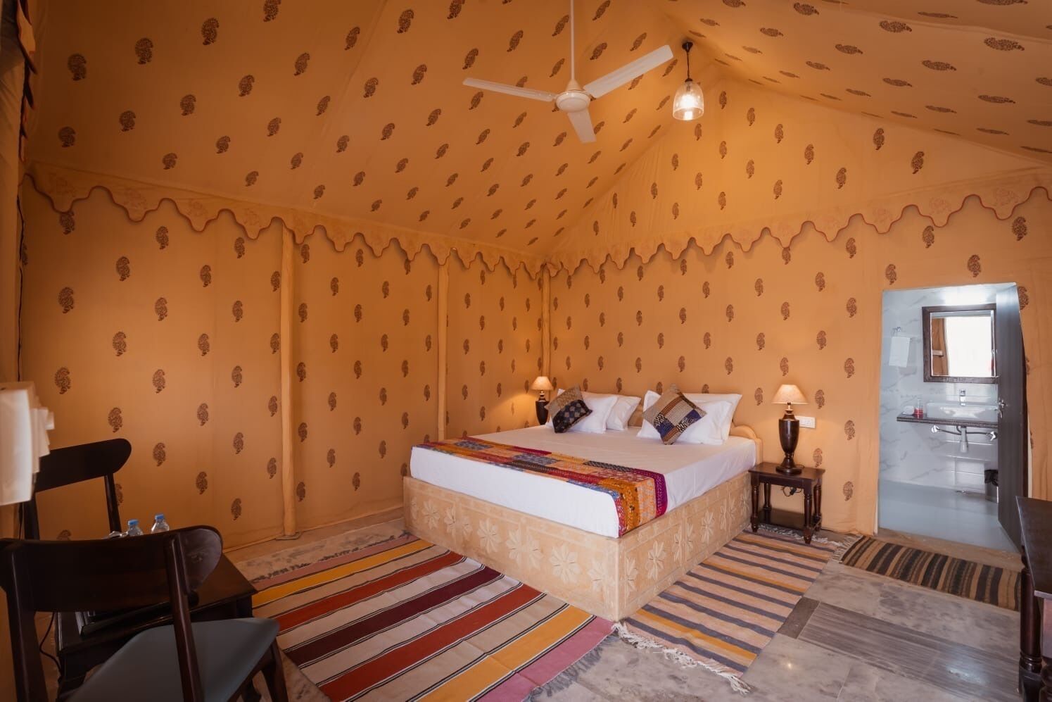 Une chambre à coucher comprenant un lit, deux tables de chevet, une chaise, un ventilateur de plafond et une salle de bain visible à travers une porte ouverte.