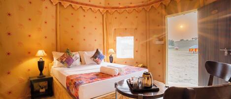 A cozy bedroom with a bed, pillows, a bedside table with a lamp, a chair, and a view of the beach.