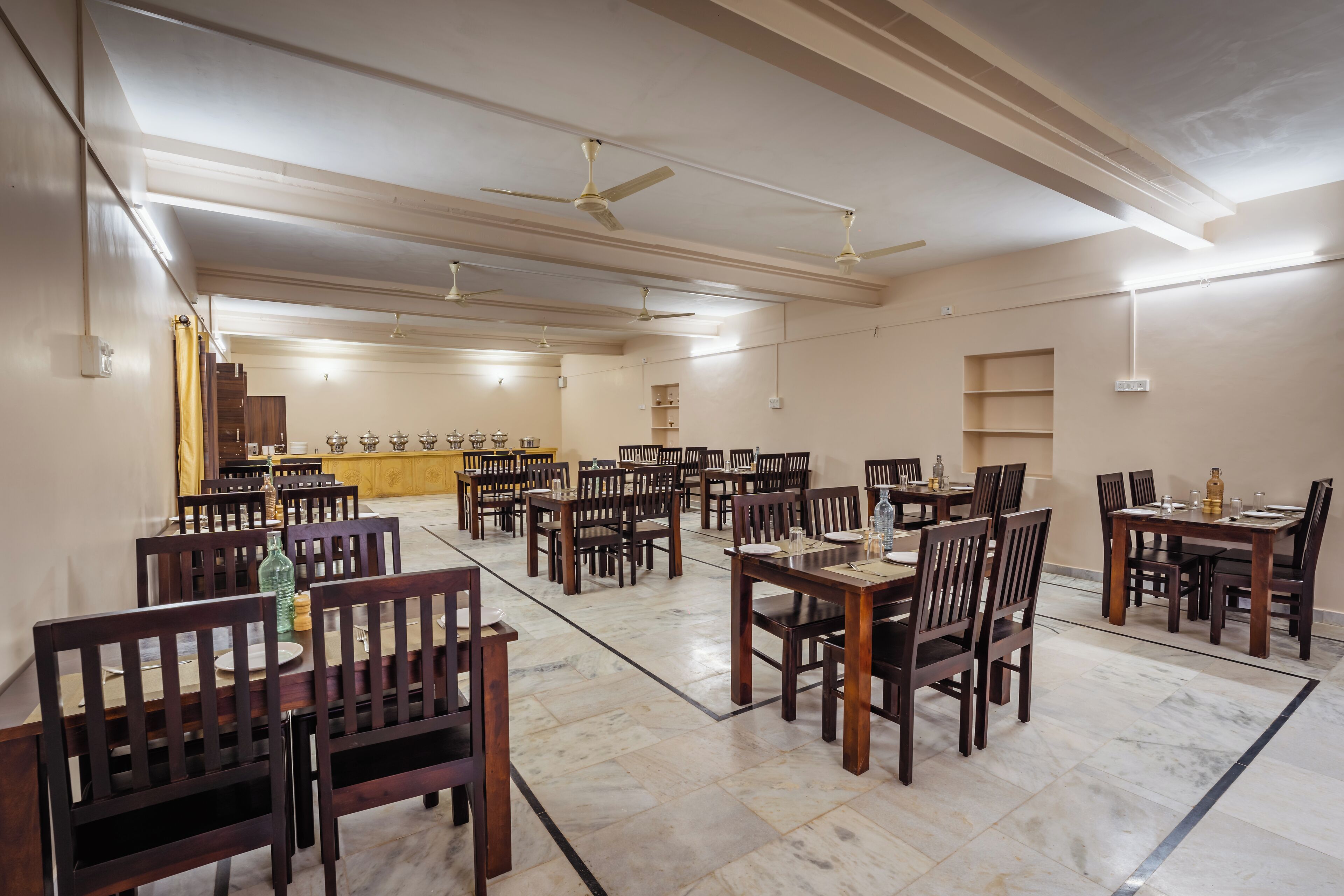 A spacious dining area with multiple tables and chairs, ceiling fans, and a kitchen area in the background.
