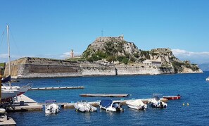 A coastal marina with boats docked, a lighthouse, and a fort on a hill.