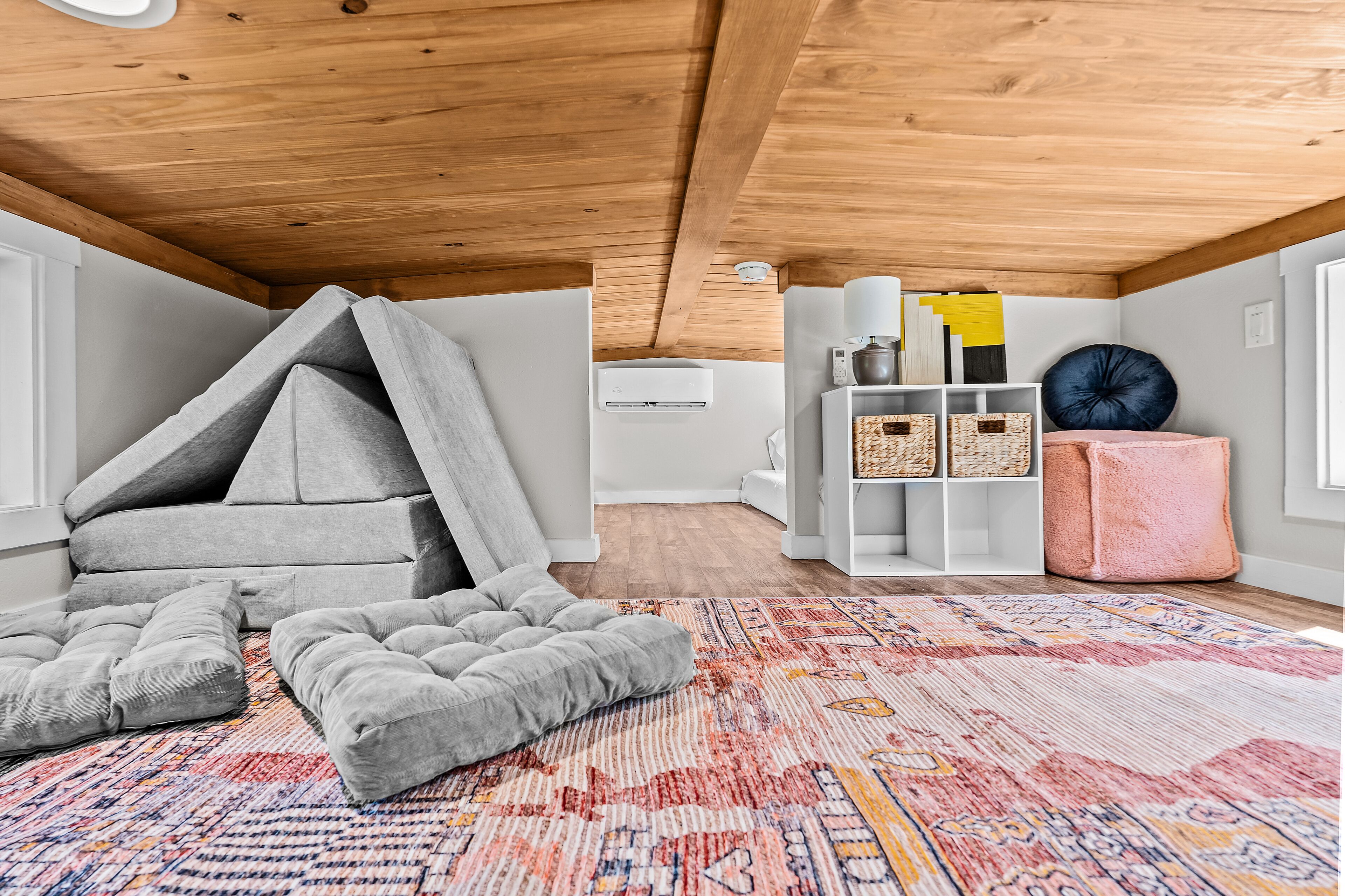 A loft bedroom with a wooden ceiling, a patterned rug, and a white shelving unit with decorative items.