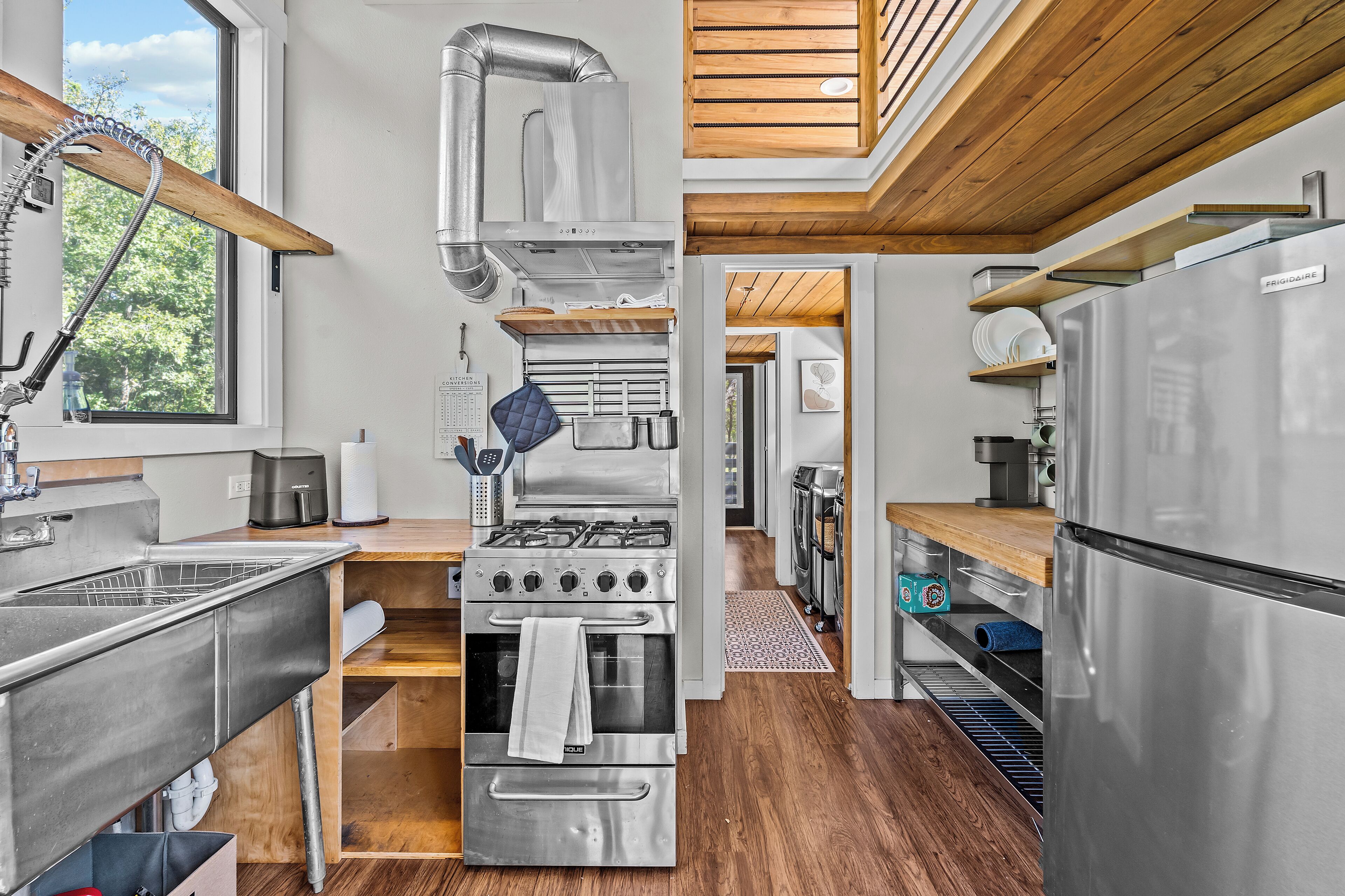 A modern kitchen with stainless steel appliances, wooden flooring, and a view of the outdoors through a large window.