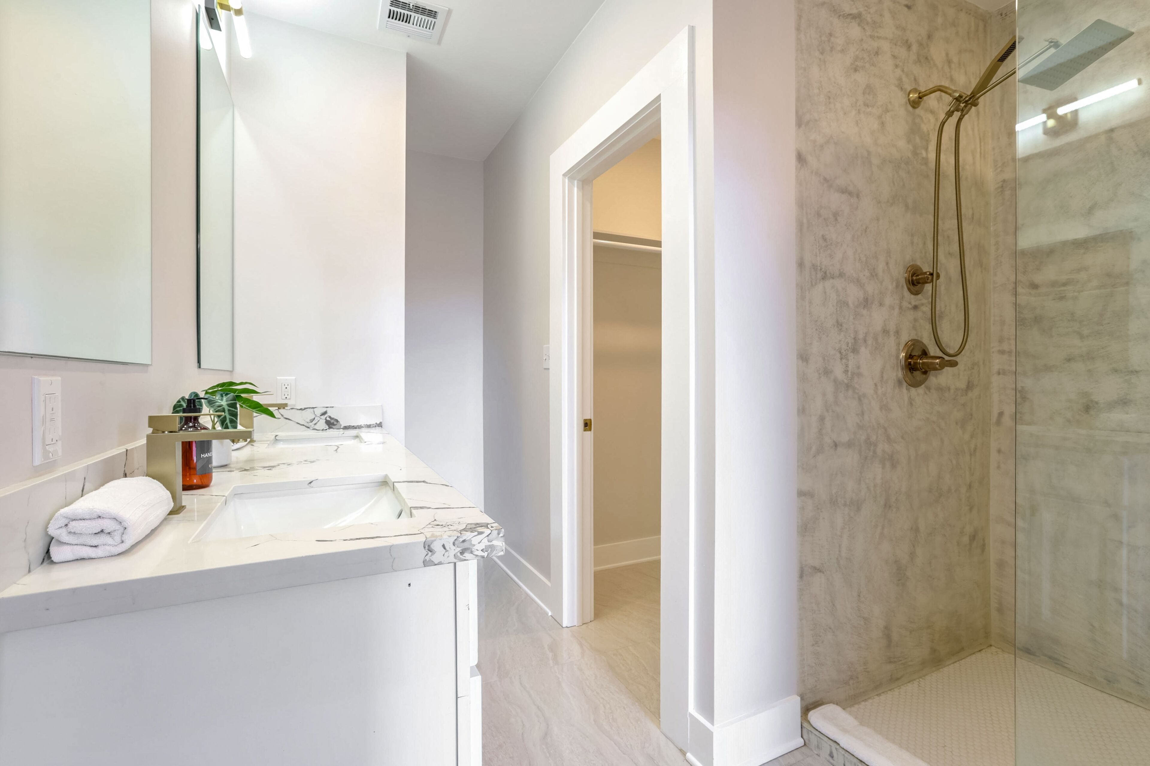 A bathroom with a large mirror, two sinks, a shower with a glass door, and a hallway leading to another room.