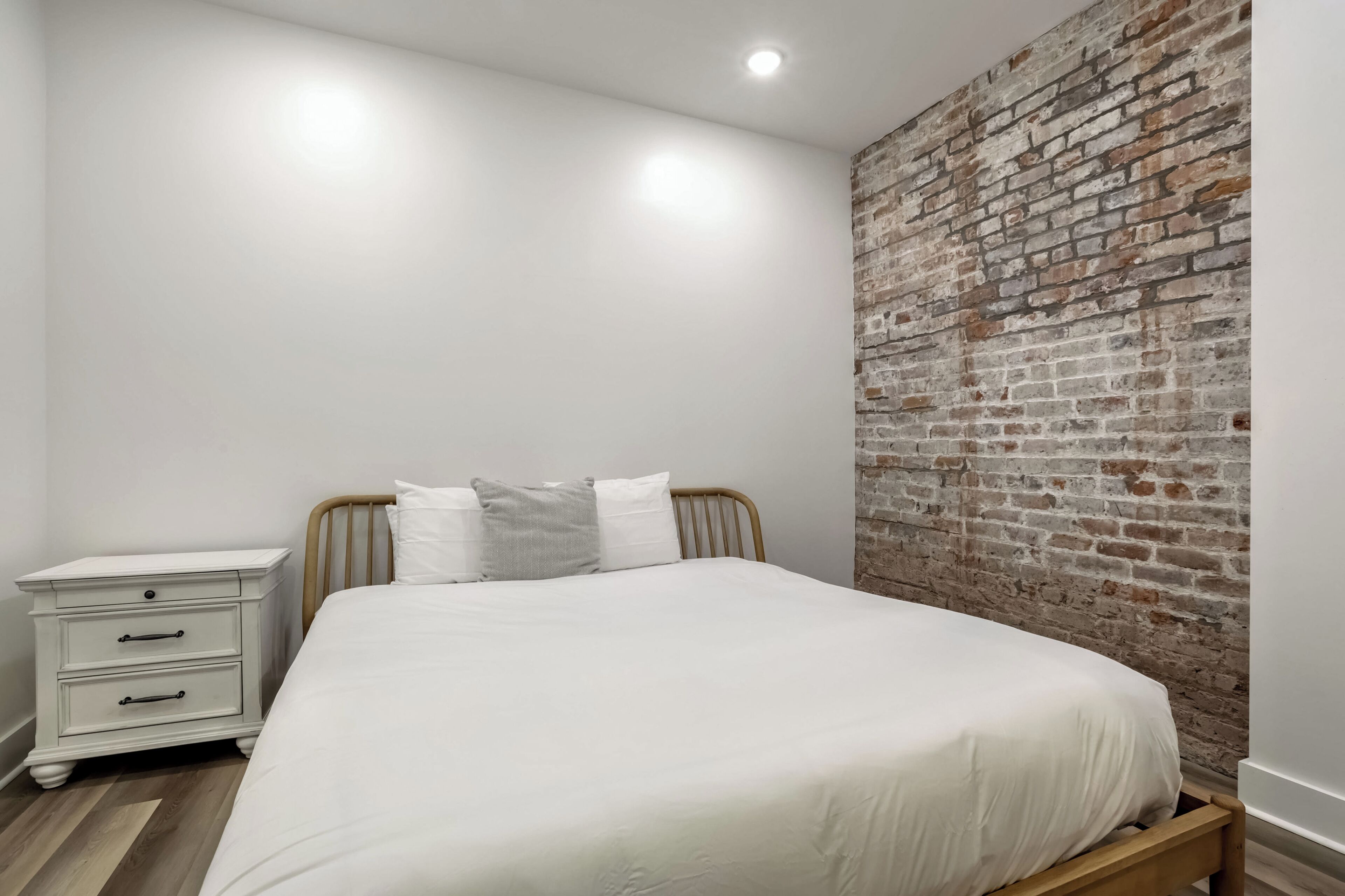 Une chambre avec un lit blanc, une table de chevet avec des tiroirs et un mur en briques.