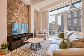 A modern living room with a brick wall, a flat-screen TV, a fireplace, a sofa, and a large window with a view of buildings. - Roami at The Churchill (New Orleans)