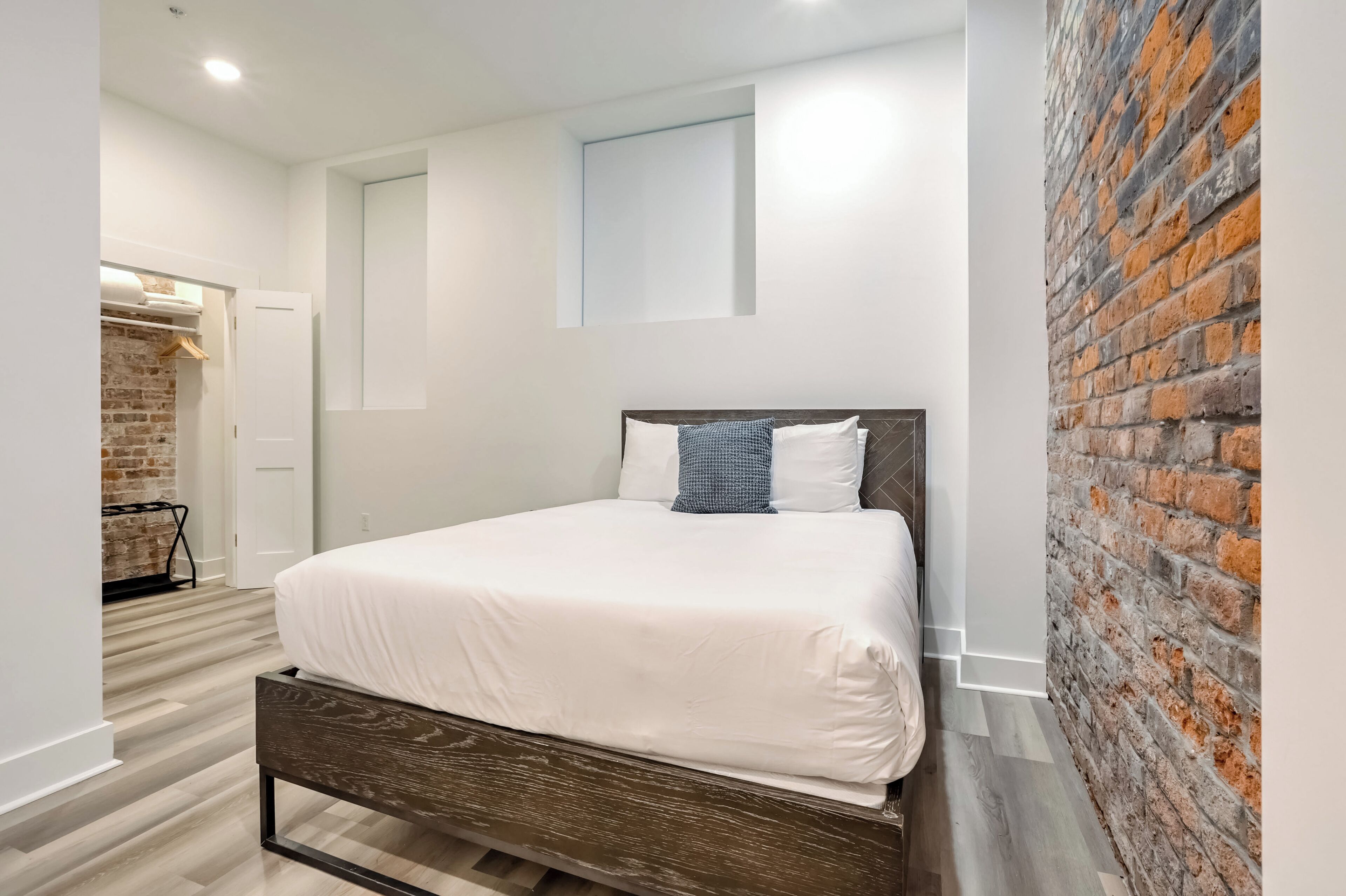 A modern bedroom with a wooden bed, white bedding, a brick accent wall, and a small window.