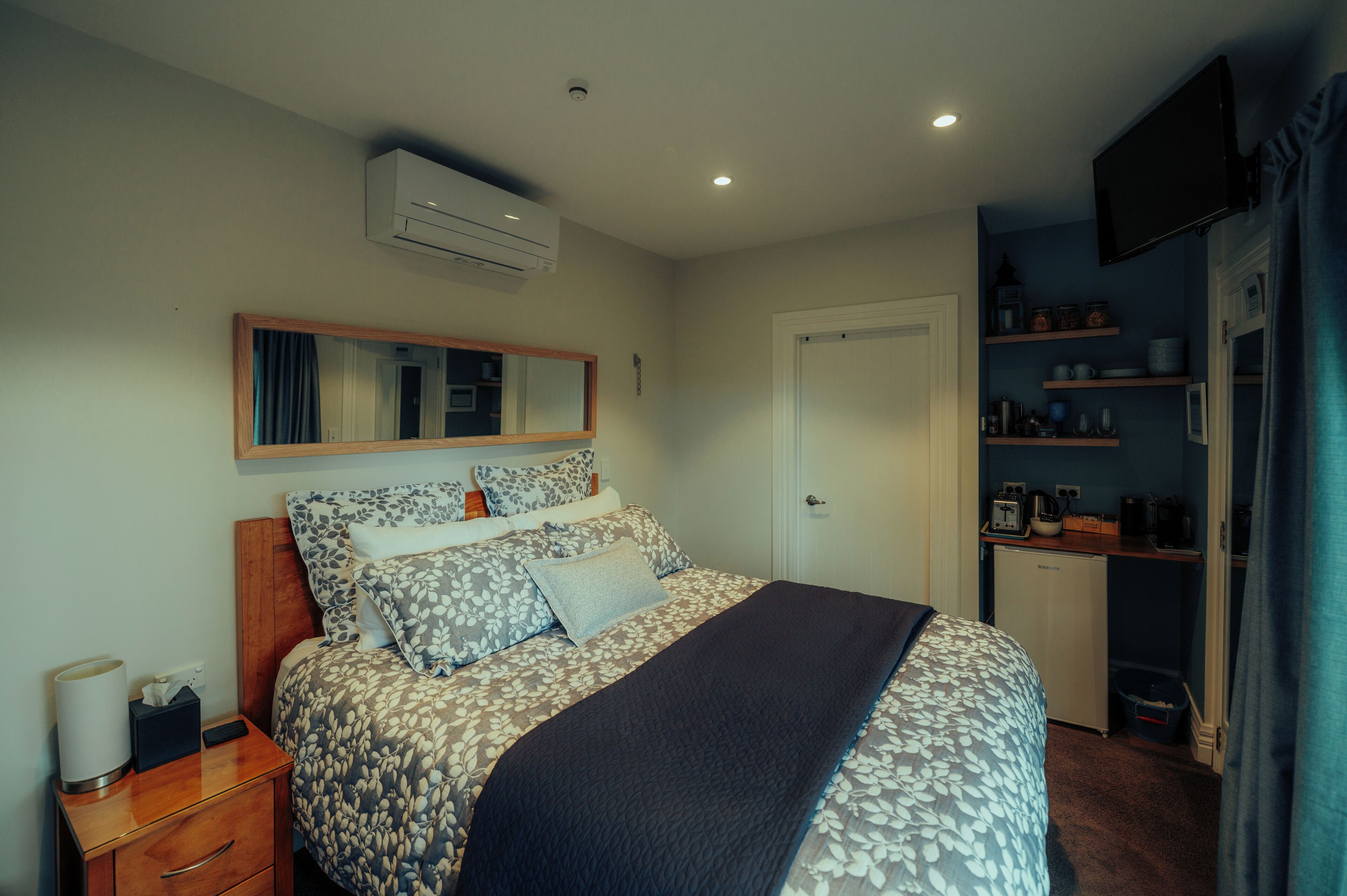 Un dormitorio con cama, mesita de noche, espejo, armario y una pequeña cocina.
