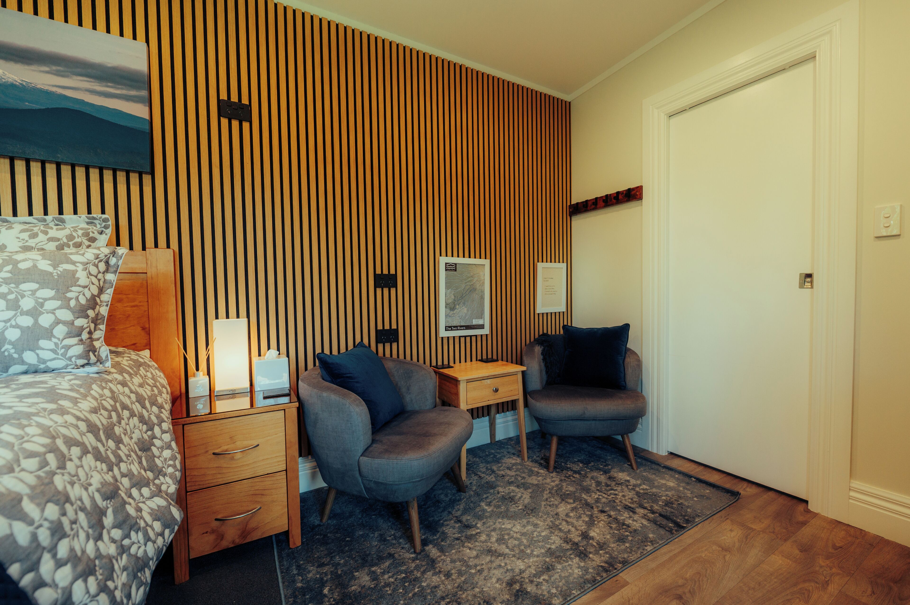 Habitación de hotel con cama, dos sillones, una mesita, una mesita de noche, un panel de madera con listones y una puerta.
