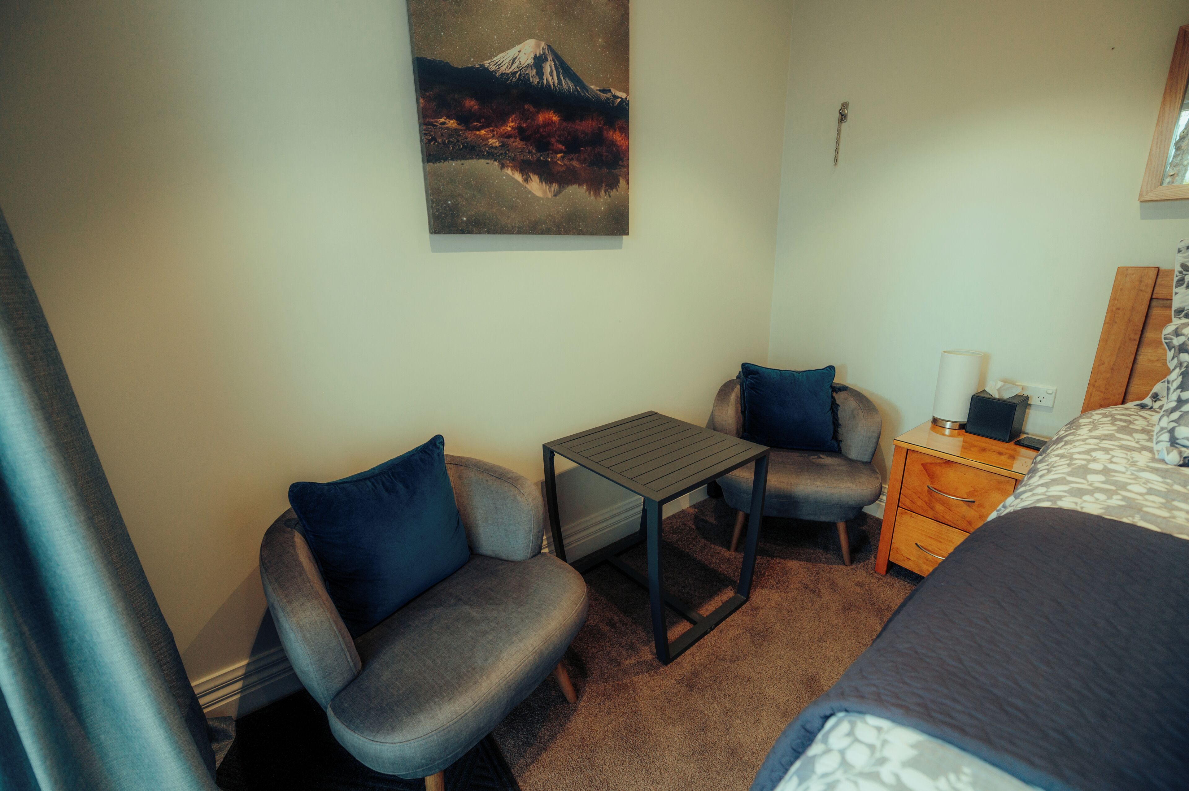 A hotel room with a bed, a small table, two armchairs, a dresser, and a painting of a mountain.