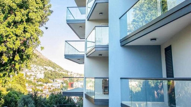 A multi-story building with balconies, large windows, and a view of a hillside.