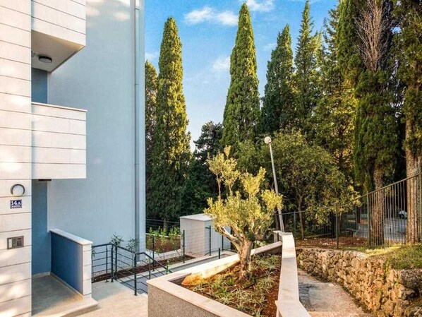 A modern building with a blue facade, a stone retaining wall, and a landscaped garden with tall trees and a small tree.
