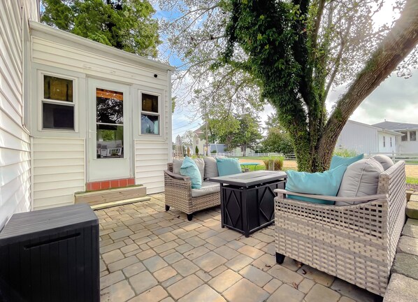 A patio area with wicker furniture and a small table, a white mobile home, and a tree in the background. - Cove Point Beach Cottage with Chesapeake Bay Views (Lusby)