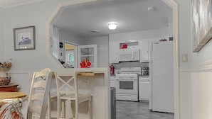 A kitchen with white cabinets, a microwave, an oven, and a refrigerator. There is a dining area with a table and chairs. - Cove Point Beach Cottage with Chesapeake Bay Views (Lusby)