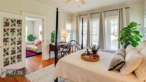 Um quarto com cama, ventilador de teto, janela com cortinas, uma planta e uma porta com desenho floral.
